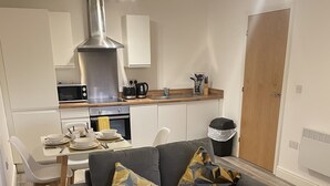 A modern living room with a grey sofa, two patterned cushions, a dining table with chairs, a kitchen area with white cabinets, a stainless steel range hood, and a wooden door. - Modern city centre two bedroom fully equipped apartment (Derby)