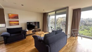 A modern living room with a flat-screen TV, two sofas, a coffee table, and a balcony with a view. - Torquay285 King Studio Beachfront Resort (Torquay)