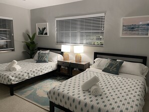 A hotel room with two beds, a nightstand with lamps, a large window with blinds, a plant, and a beach-themed pillow.