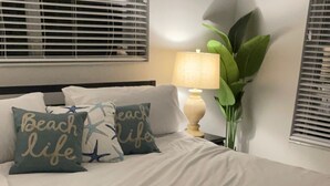 Ein ordentlich bezogenes Bett mit weißen Leinen und einem aufgerollten Handtuch, zwei Kissen mit dem Aufdruck 'Beach Life', eine Lampe und eine Pflanze.