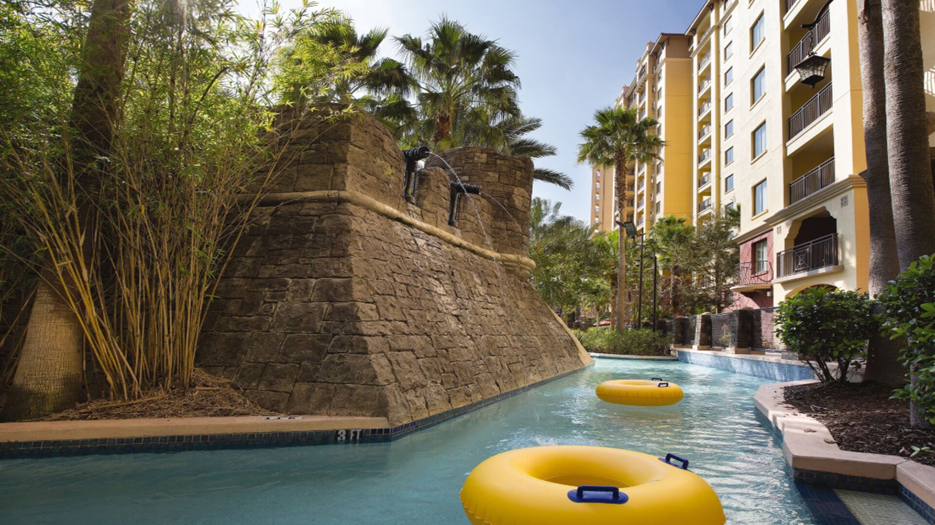 A pool area with a stone wall, palm trees, and a yellow building in the background.