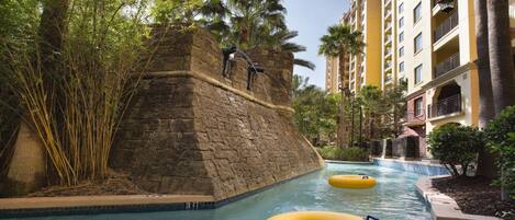 A pool area with a stone wall, palm trees, and a yellow building in the background.