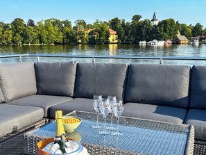 A boat deck with a cushioned seating area, a table with a bottle of champagne, glasses, and a bowl of snacks.