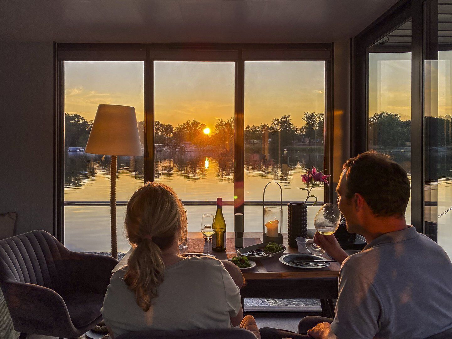 Un coin repas offrant une vue sur un coucher de soleil sur l’eau, avec une table agrémentée d’une bouteille, d’une bougie et de verres.