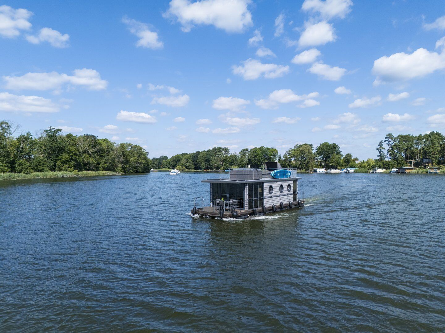Una casa flotante en un lago tranquilo, con un bote amarrado al fondo y árboles a lo largo de la orilla.