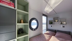 A bedroom with a bed, a circular window, and built-in shelves with various items. - Houseboat Suite GRAND PLAISIR – Luxury houseboat, no license required (Zehdenick)