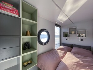 Une chambre à coucher avec un lit, une fenêtre ronde et des étagères intégrées où sont rangés divers objets.