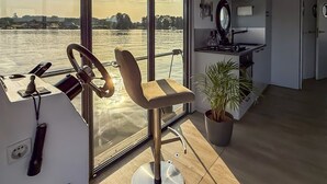 A modern room with a large window overlooking a body of water, a steering wheel, a chair, a potted plant, and a kitchen area. - Houseboat Suite GRAND PLAISIR – Luxury houseboat, no license required (Zehdenick)