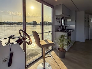 Une pièce moderne avec une grande fenêtre donnant sur une étendue d’eau, un volant, une chaise, une plante en pot et un coin cuisine.