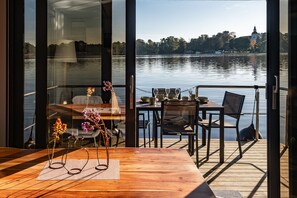 A dining area with a wooden table and chairs, overlooking a calm lake with a distant church spire.