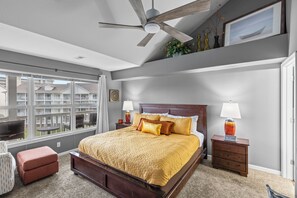 Un dormitorio con una cama grande, un ventilador de techo, una mesita de noche con una lámpara, una ventana con vistas a edificios y una pequeña mesa con sillas.