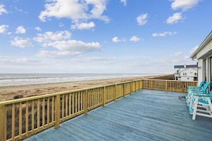 A wooden deck with a railing overlooking a beach, a few lounge chairs, and a building in the background. - Brannen's Beachfront by Ryson Vacation Rentals (Galveston)