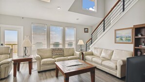 A spacious living room with two beige sofas, a wooden coffee table, and a staircase with a railing. - Brannen's Beachfront by Ryson Vacation Rentals (Galveston)
