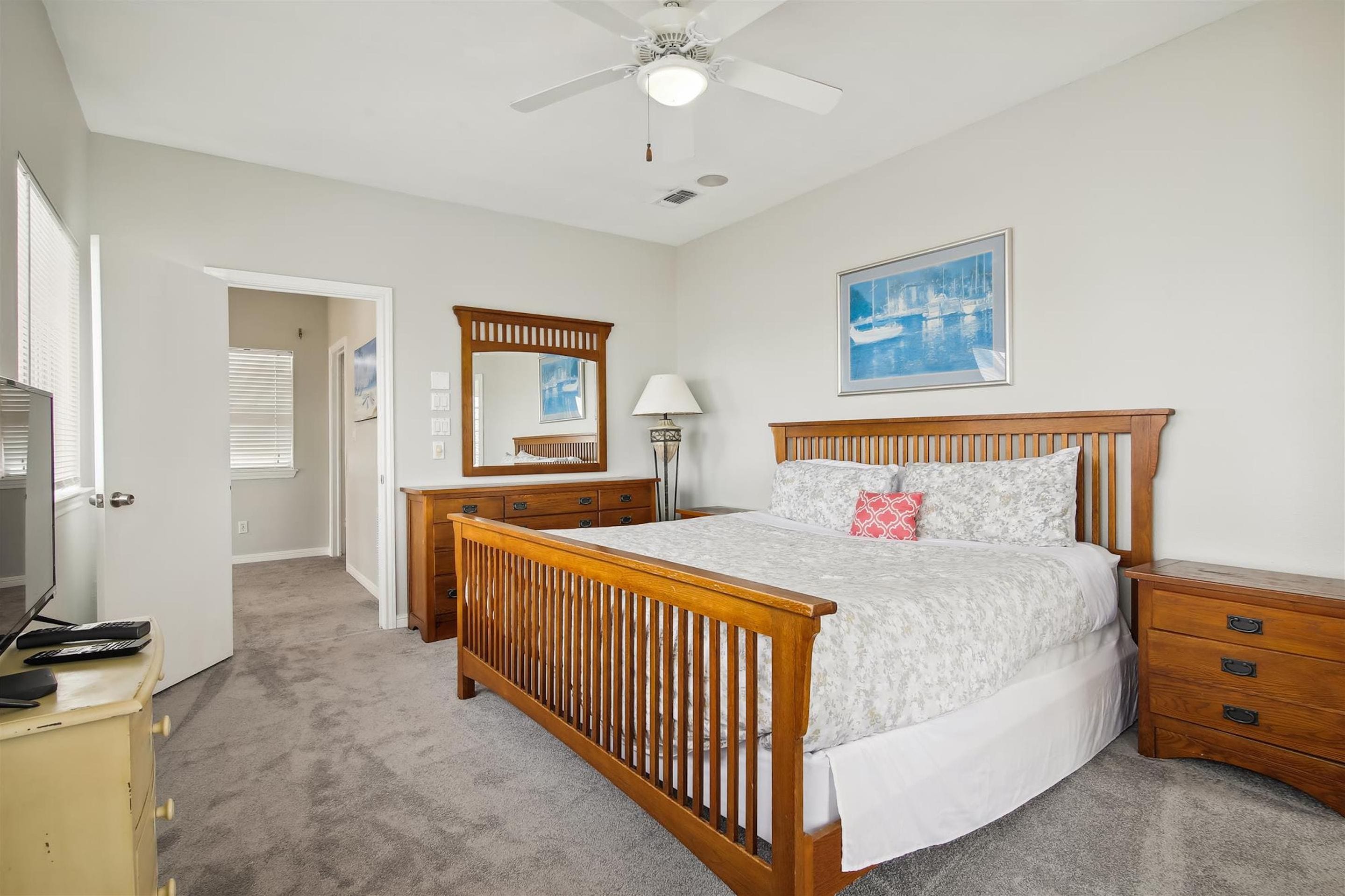 A bedroom with a wooden bed, a bedside table, a nightstand, a ceiling fan, a wall-mounted picture, and a door leading to another room.