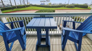 A balcony with blue chairs and a table, overlooking a parking lot with palm trees and a green lawn.