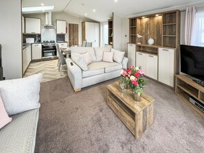 A modern living room with a grey sofa, a coffee table with flowers, a TV, and a kitchen in the background.