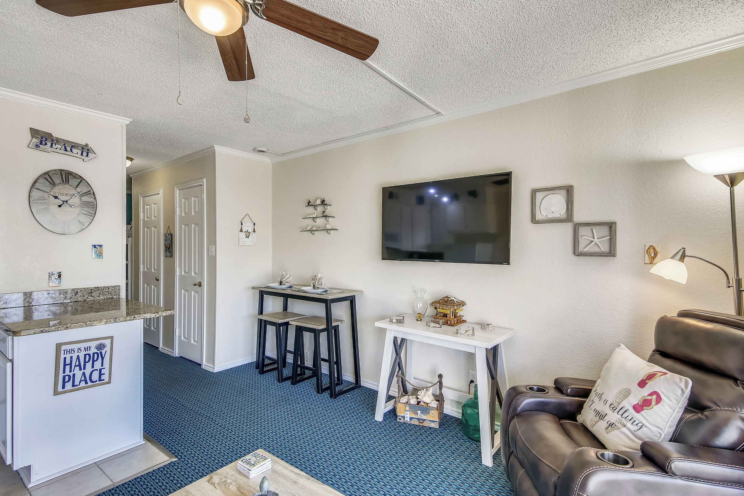 A living room with a blue carpet, a brown leather couch, a TV, a ceiling fan, and a small table with a sign that says "THIS IS MY HAPPY PLACE".