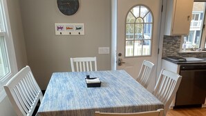 Ein Essbereich mit einem Tisch, der mit einer strukturierten Tischdecke bedeckt ist, umgeben von weißen Stühlen. An der Wand hängen eine Uhr und ein Schild mit Namen.