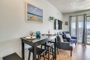 A modern living room with a dining table, two chairs, a TV mounted on the wall, and a large window with curtains. - Saltwater Paradise by Ryson Vacation Rentals (Galveston)