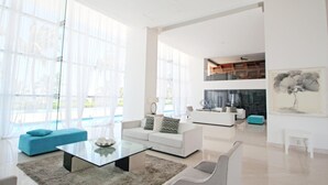 A spacious living room with a glass coffee table, white sofas, and a large window with sheer curtains.