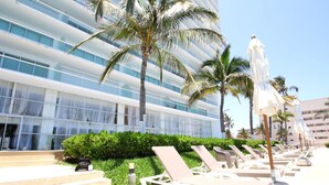 A hotel with a modern design, featuring a pool area with lounge chairs and umbrellas, surrounded by palm trees and a well-maintained lawn.