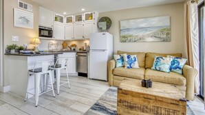 Une cuisine moderne avec des meubles blancs, un four à micro-ondes et un réfrigérateur. On y trouve un canapé beige avec des coussins bleus et une table basse en bois.