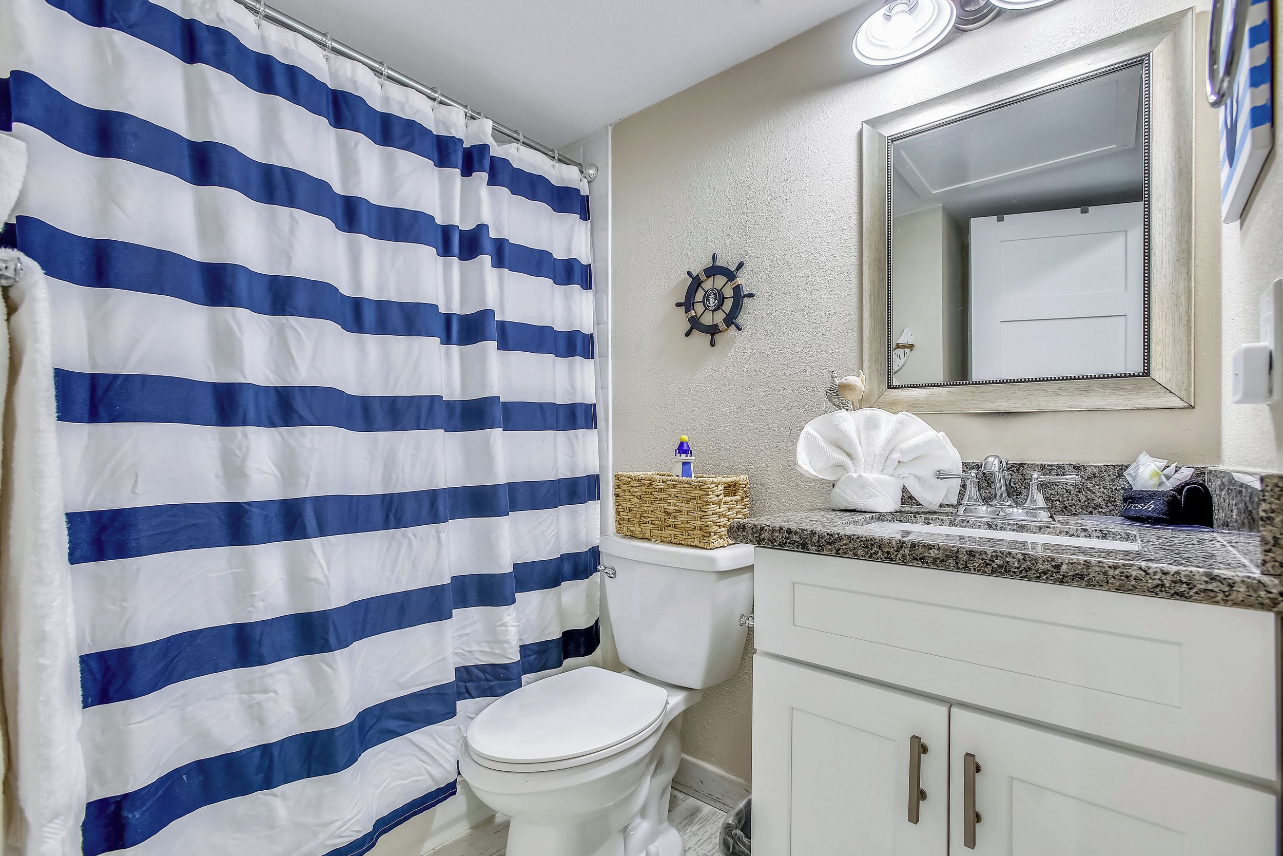 A bathroom with a striped shower curtain, a ship's wheel decoration, a toilet, and a sink with a mirror above it.