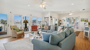 A spacious living room with a large grey sofa, wicker chairs, and a dining area with a table and chairs. - Casa Rosa West by Ryson Vacation Rentals (Galveston)