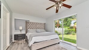 Una camera da letto con un letto grande, un ventilatore a soffitto, un comodino, una lampada e una porta scorrevole in vetro che si apre su un giardino.