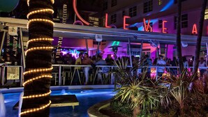 Esterno di un hotel ben illuminato con insegne al neon, area piscina e posti a sedere all'aperto.