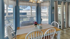 A dining area with a wooden table and white chairs, large windows with blinds, and a view of buildings outside. - Catch of the Day by Ryson Vacation Rentals (Galveston)