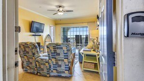 A room with a ceiling fan, a television, a window, and a table with a lamp. - Dawn's Early Light by Ryson Vacation Rentals (Galveston)