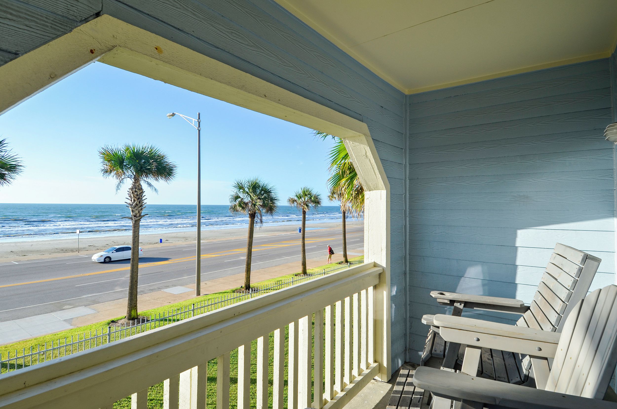 Un balcón con vista a la playa y al océano, con barandilla blanca y una silla de madera.