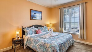 A bedroom with a patterned bedspread, a standing lamp, a ceiling fan, a window with blinds, and a framed picture on the wall. - Dawn's Early Light by Ryson Vacation Rentals (Galveston)