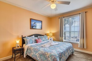 A bedroom with a patterned bedspread, a standing lamp, a ceiling fan, a window with blinds, and a framed picture on the wall. - Dawn's Early Light by Ryson Vacation Rentals (Galveston)