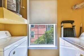 A laundry room with two white washers and dryers, a window with a view of a red building, and a shelf with a newspaper.