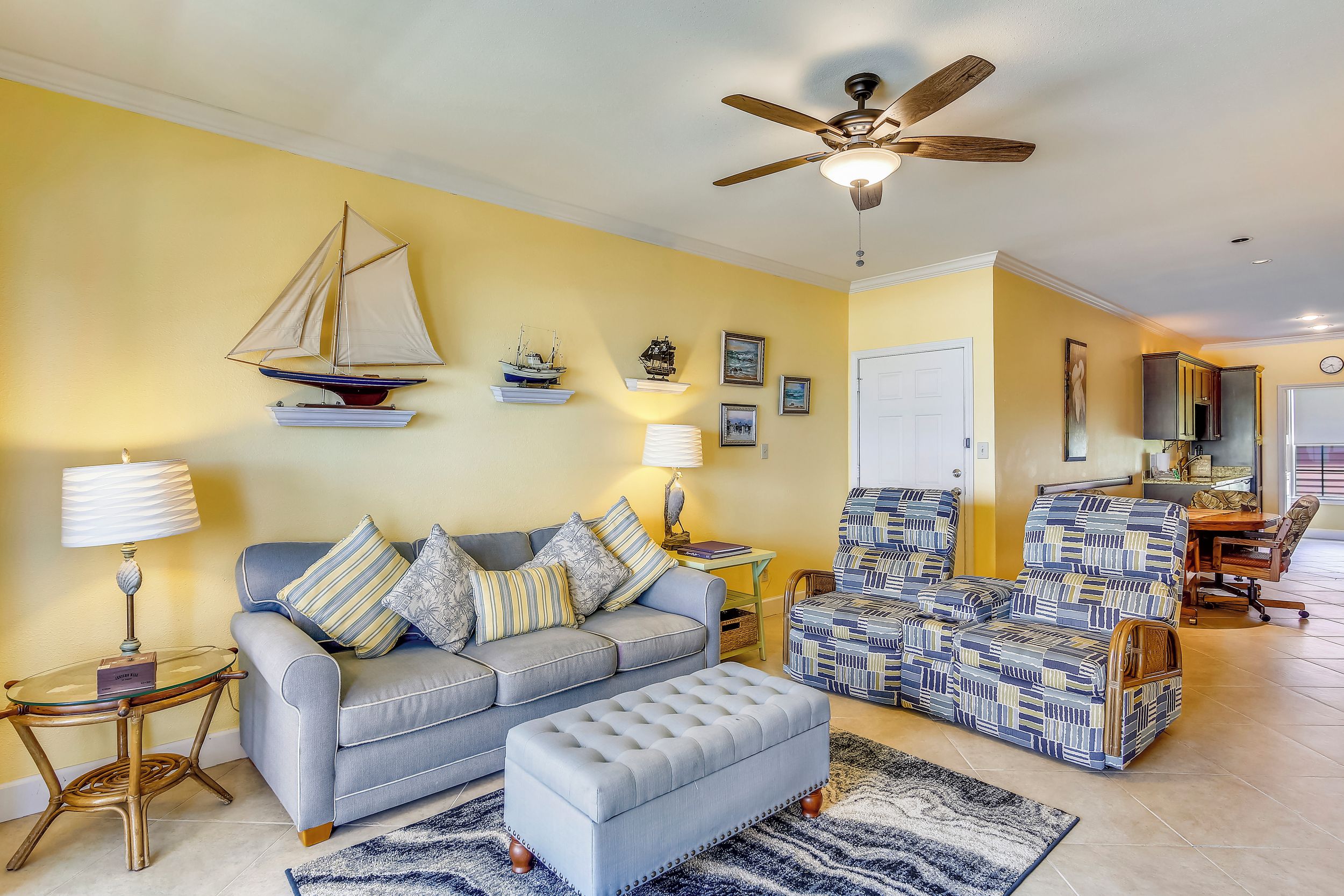Una sala de estar con sofá, reposapiés y sillón estampado, un ventilador de techo y estantes fijos en la pared con objetos decorativos.