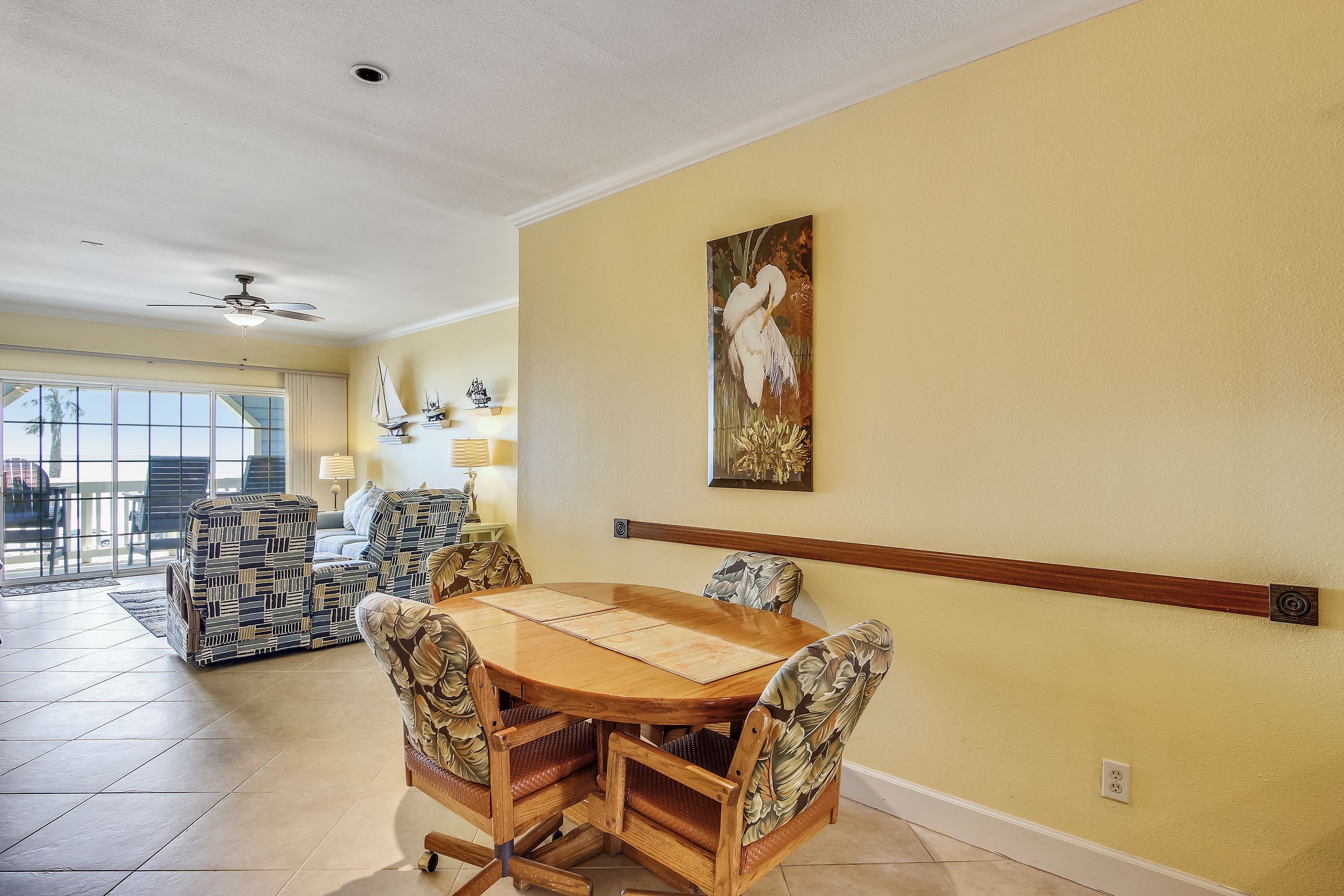 A dining area with a wooden table and chairs, a ceiling fan, and a large window with a view of palm trees.