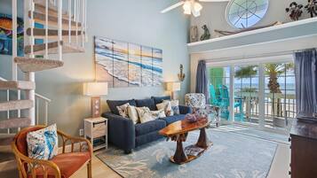A living room with a blue sofa, a wooden coffee table, a rattan chair, a staircase with a cat tree, a painting of a beach, and a view of the ocean through large windows.
