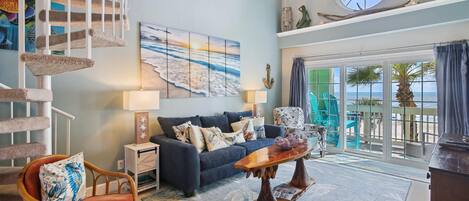 A living room with a blue sofa, a wooden coffee table, a rattan chair, a staircase with a cat tree, a painting of a beach, and a view of the ocean through large windows.