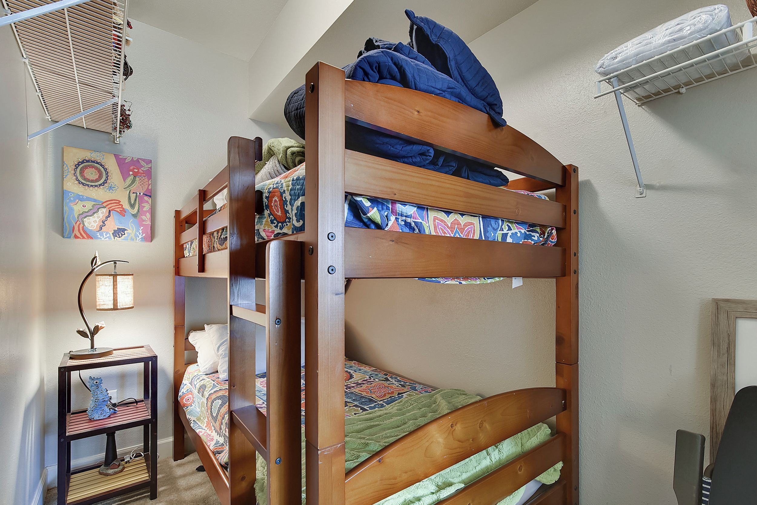 Ein Stockbett mit Holzrahmen, ein kleiner Tisch mit Lampe und ein an der Wand befestigtes Regal.