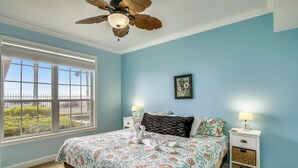 A bedroom with a bed, two bedside tables, a ceiling fan, a window with blinds, and a view of greenery outside. - Turtle Reef by Ryson Vacation Rentals (Galveston)
