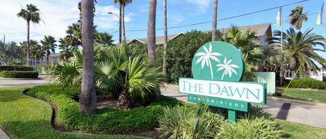 A sign for The Dawn Condominiums with palm trees and a well-maintained lawn.