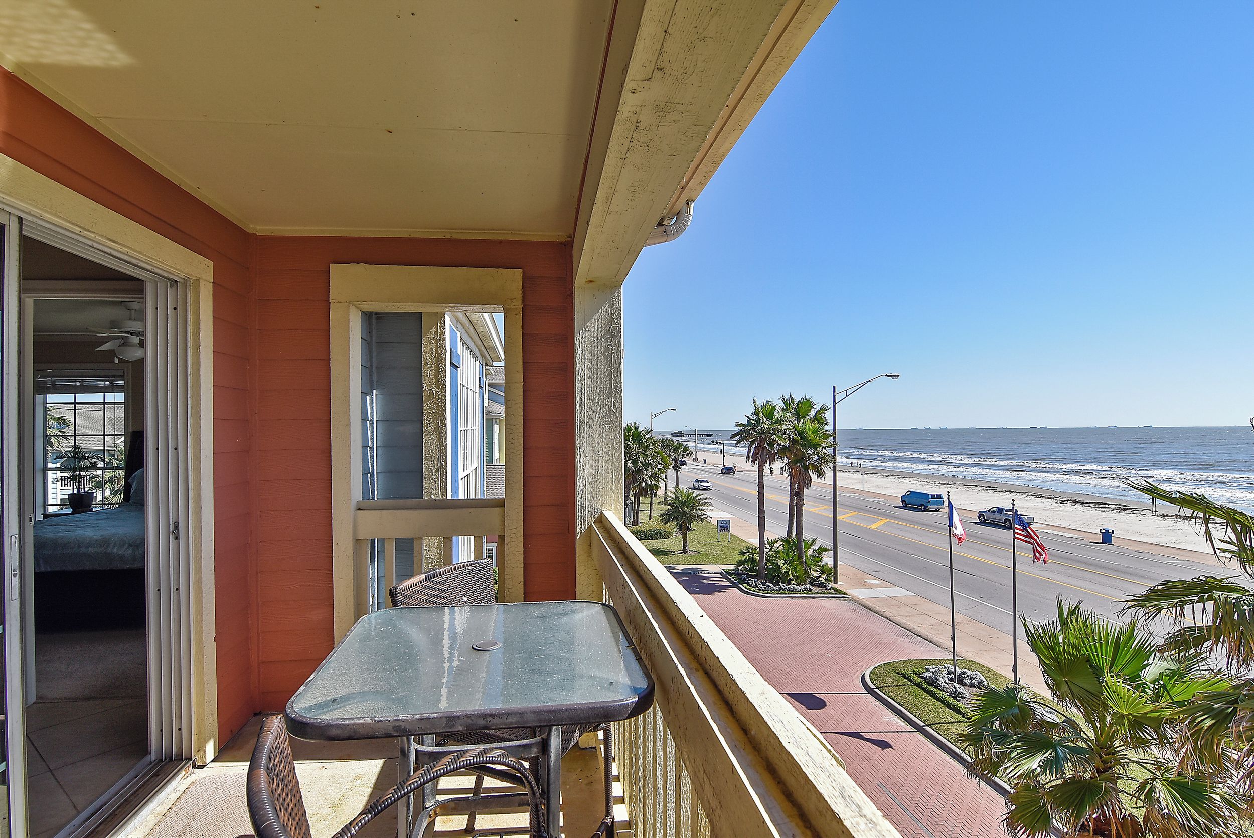 Een balkon met een tafel en stoelen, met uitzicht op een strand met palmbomen en een weg met auto's.