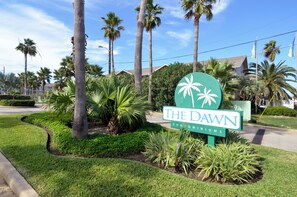 A sign for The Dawn Condominiums with palm trees and a well-maintained lawn.