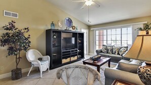A living room with a ceiling fan, a large window, a sofa, a chair, and a bookshelf. - Dawn 631 by Ryson Vacation Rentals (Galveston)
