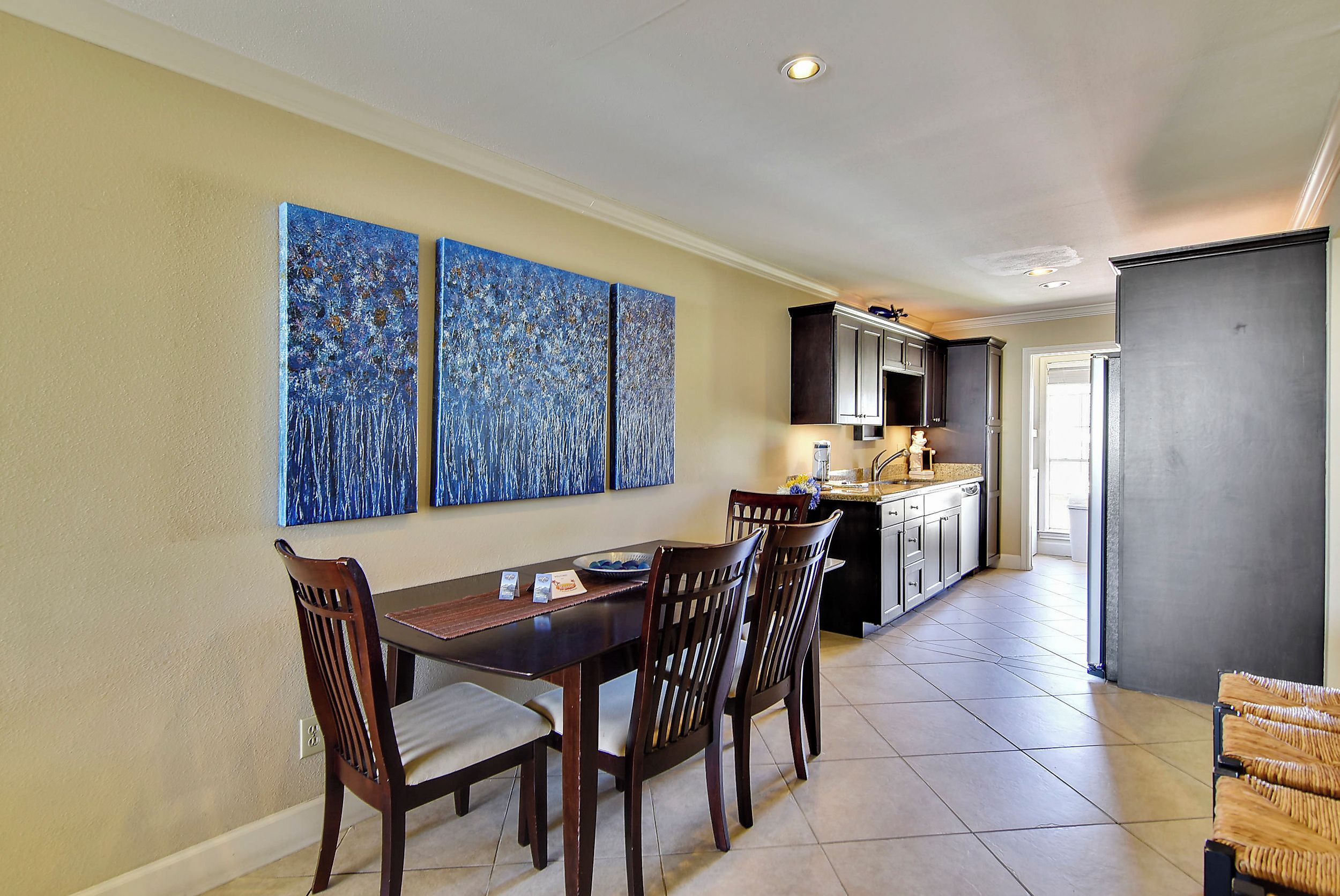 Un coin repas avec une table et des chaises en bois, une cuisine en arrière-plan et trois tableaux abstraits bleus accrochés au mur.