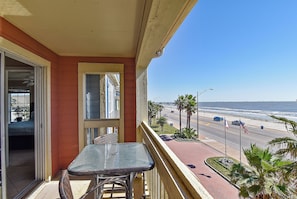 A balcony with a table and chairs, overlooking a beach with palm trees and a road with cars.