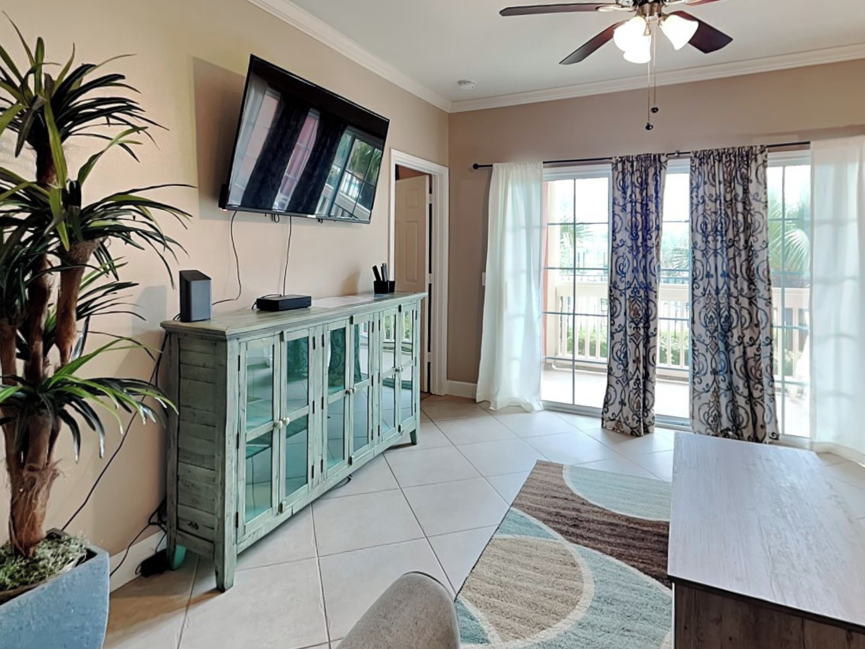 A living room with a flat-screen TV mounted on the wall, a glass cabinet with drawers, a ceiling fan, and a large window with curtains.