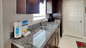 A kitchen with granite countertops, a double sink, dark wooden cabinets, and a white door. - Just for Fun by Ryson Vacation Rentals (Galveston)